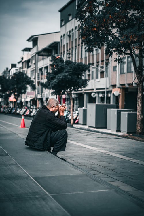 A Man in Black Jacket Sitting on Sidewalk