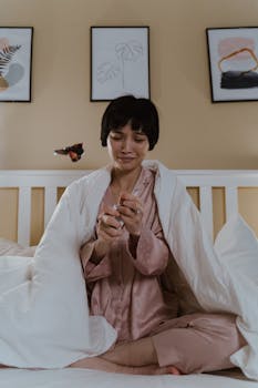 Asian woman in pajamas feeling upset in a bedroom, wrapped in a blanket.
