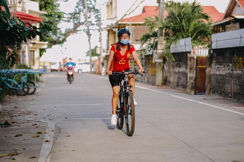 Free A woman in a red shirt rides a bike on a quiet urban street, enjoying a leisurely afternoon. Stock Photo