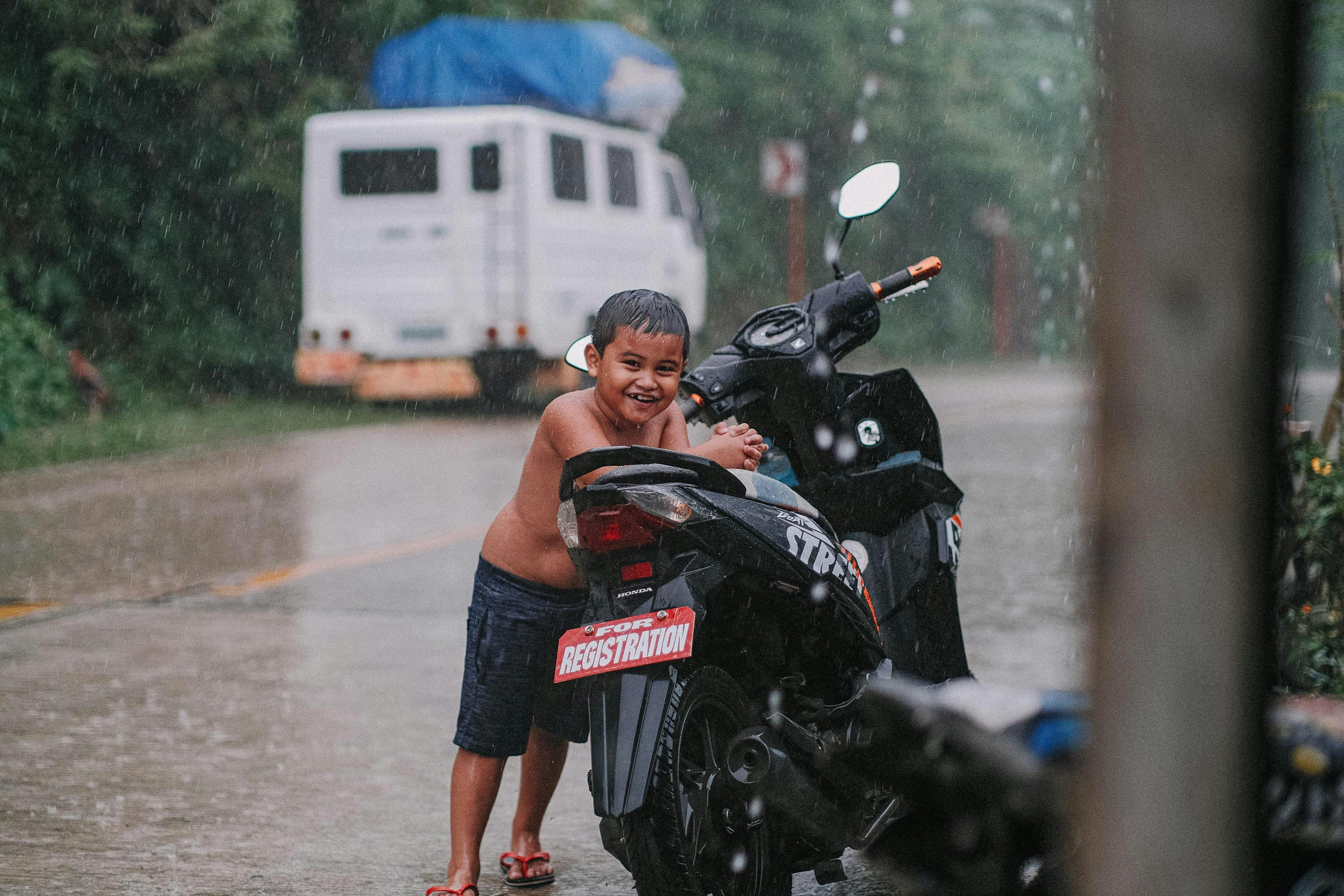 a shirtless boy standing beside a motorcycle under the rain