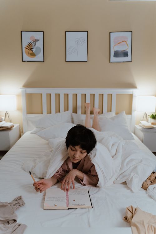 Free Asian woman lying on bed indoors, enjoying a relaxed moment with a notebook. Cozy at-home setting. Stock Photo