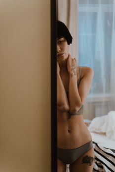 Artistic image of a tattooed woman in lingerie leaning against a mirror indoors.