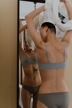 Young woman adjusting shirt in front of mirror, showcasing lingerie and reflection.