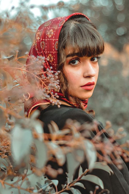 Free A beautiful woman with bangs wearing a red headscarf gazes at the camera amidst foliage. Stock Photo