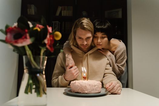Couple shares a heartfelt moment around a birthday cake with lit candles indoors.