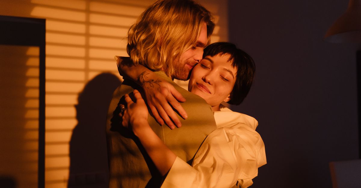 An affectionate couple shares a warm embrace in soft evening light by window blinds.