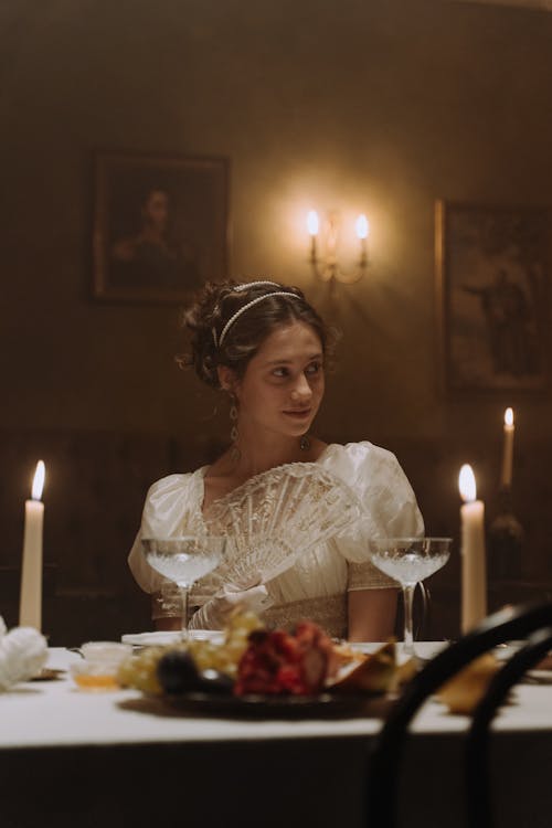 Woman in White Dress Sitting Holding Hand fan Sitting at Table with Drinks