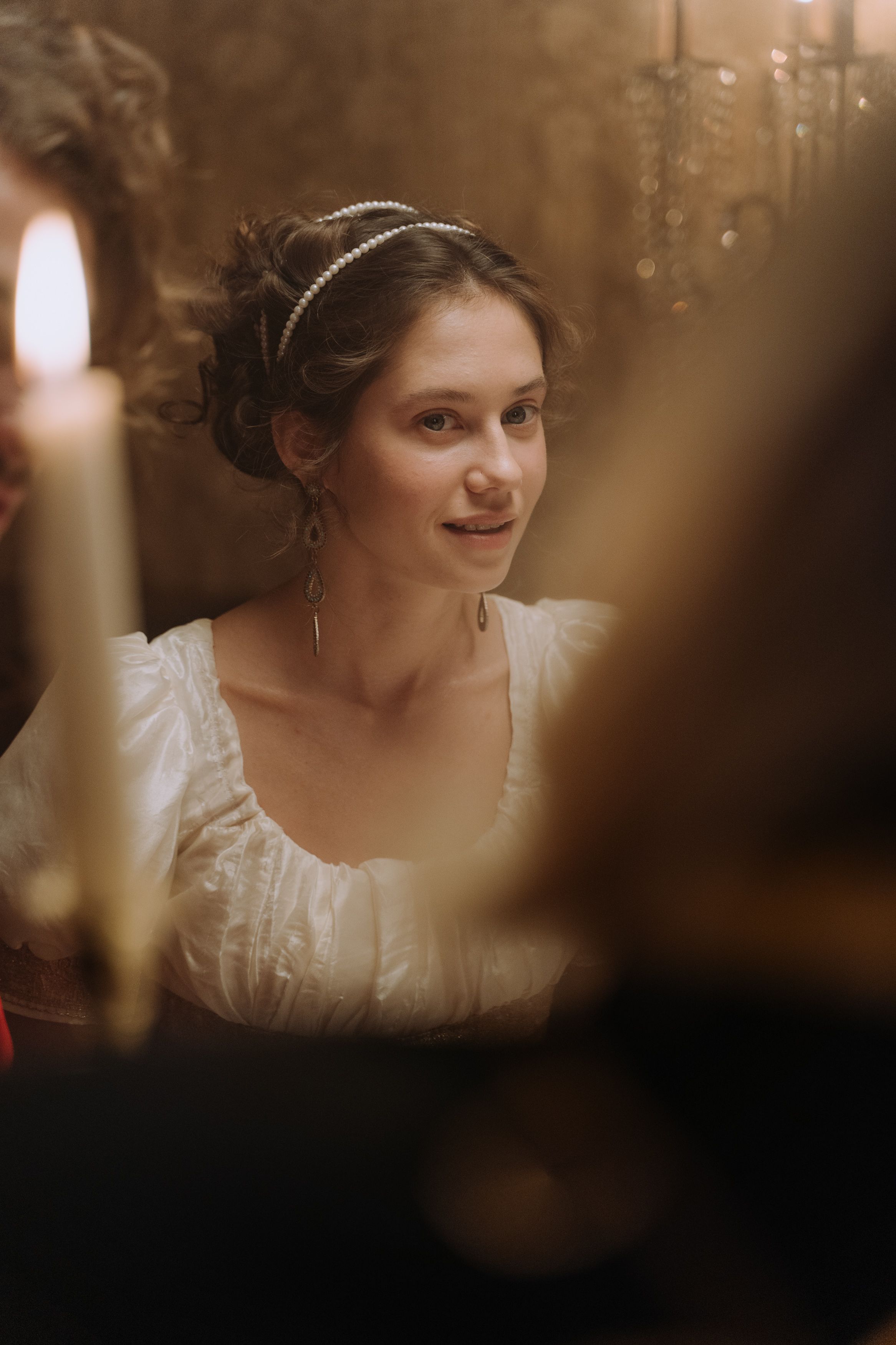 A woman in vintage attire sits elegantly under warm candlelight, evoking a classic, timeless era.