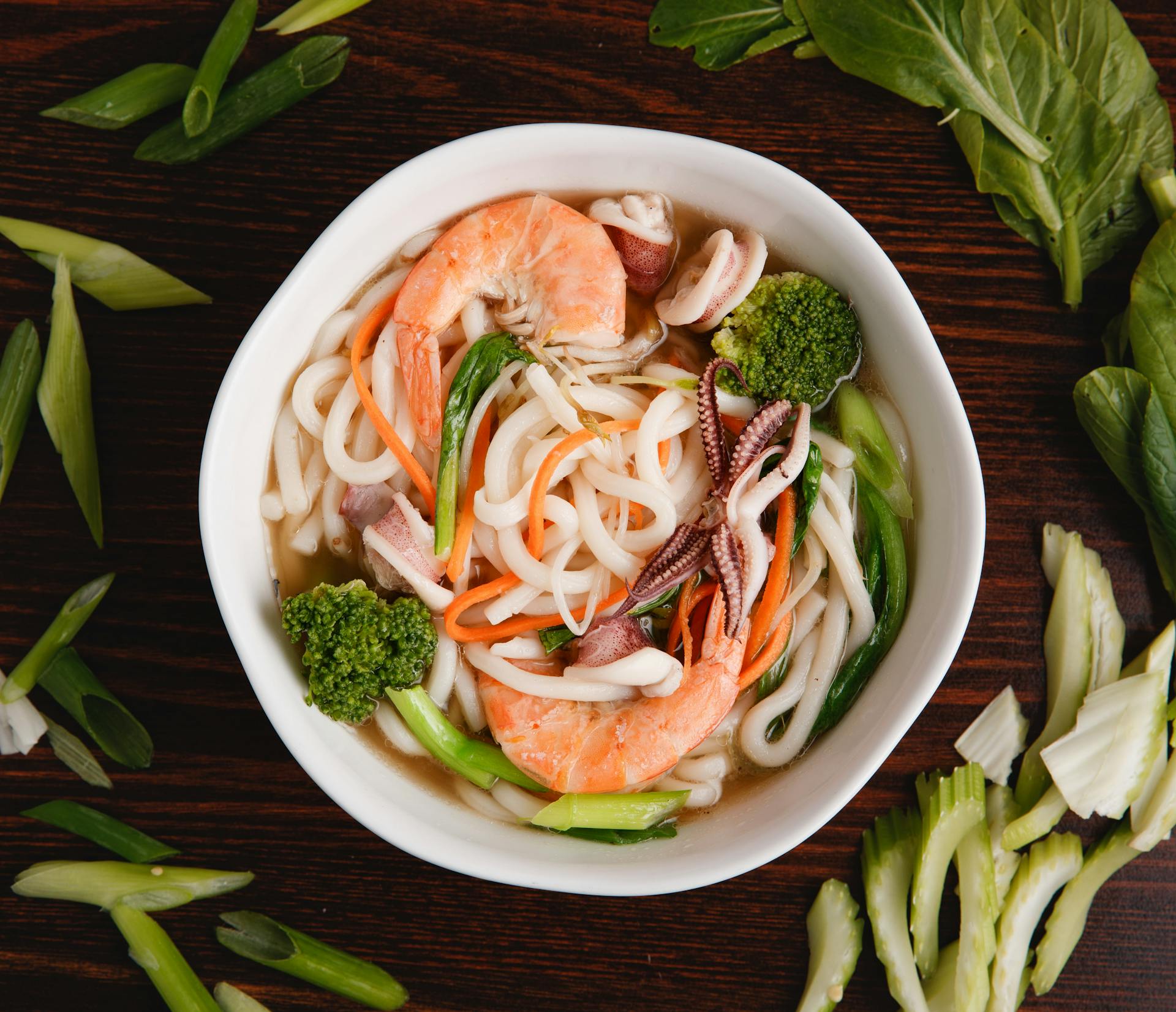 Delicious Asian udon noodle soup topped with prawns, squid, and fresh vegetables in a white bowl.