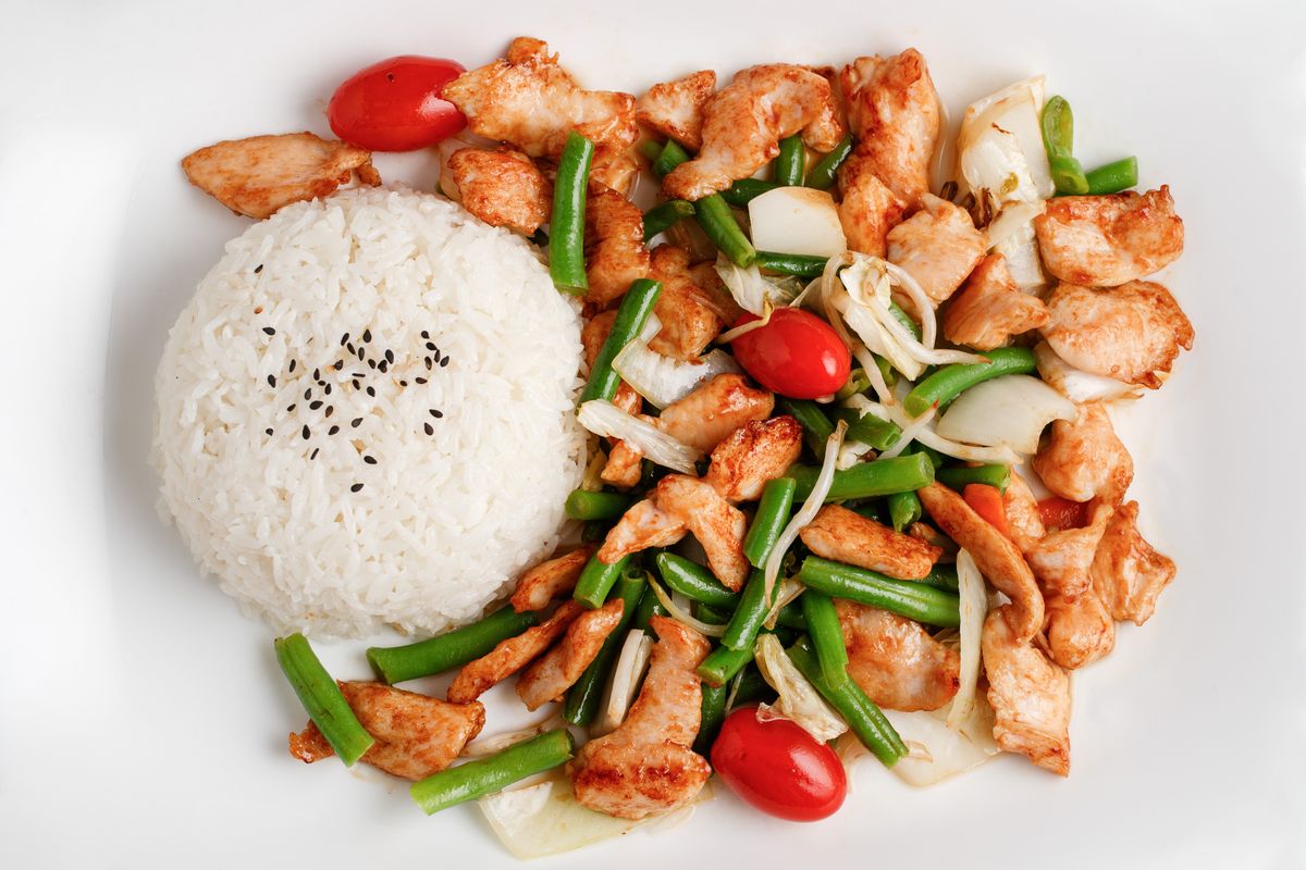 Stir-fried chicken pieces with green beans, onion and cherry tomatoes beside rice with black sesame