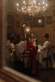 A group of elegantly dressed adults dining in a vintage room with a chandelier, captured through a window.