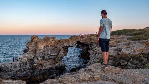 Free A man stands on a rocky cliff at sunset, gazing at the ocean and natural arch formations. Stock Photo