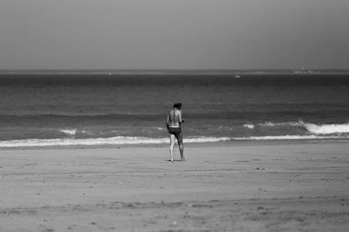 A Grayscale Photo of a Shirtless Man Walking on the Beach
