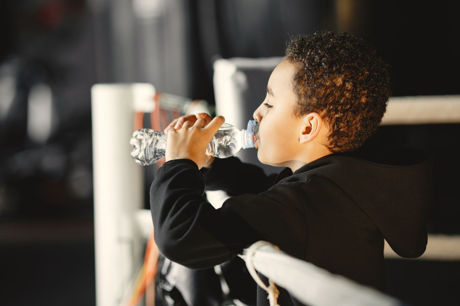 Side view of a young boy in a black hoodie hydrating with a water bottle inside a boxing gym.