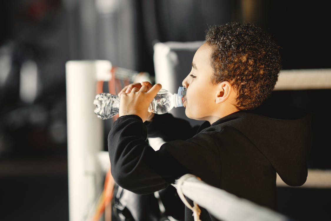 Free Side view of a young boy in a black hoodie hydrating with a water bottle inside a boxing gym. Stock Photo