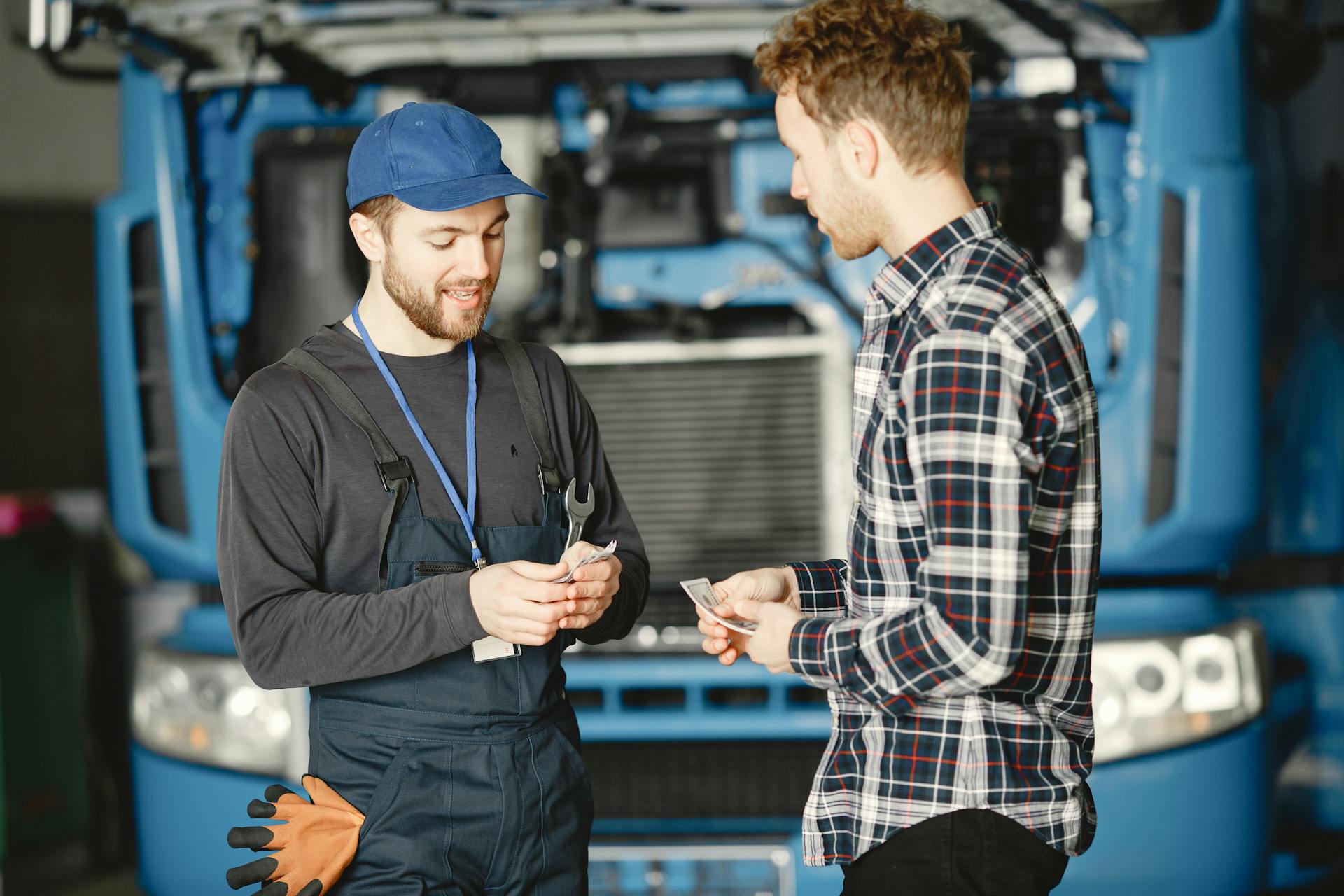 Auto mechanic in blue uniform receives payment from customer in a garage setting with a truck in the background.