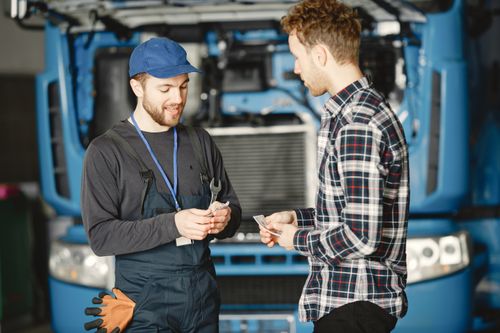 Free Auto mechanic in blue uniform receives payment from customer in a garage setting with a truck in the background. Stock Photo