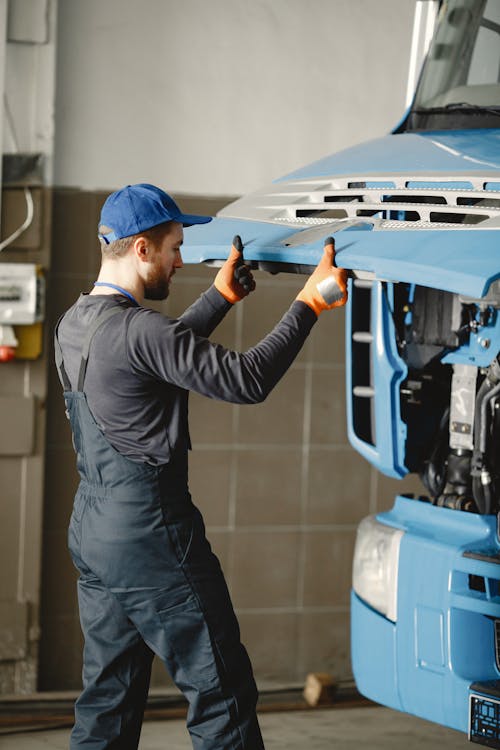 Free Man in Work Clothes and Blue Cap Opening Front Mask of Blue Truck Stock Photo