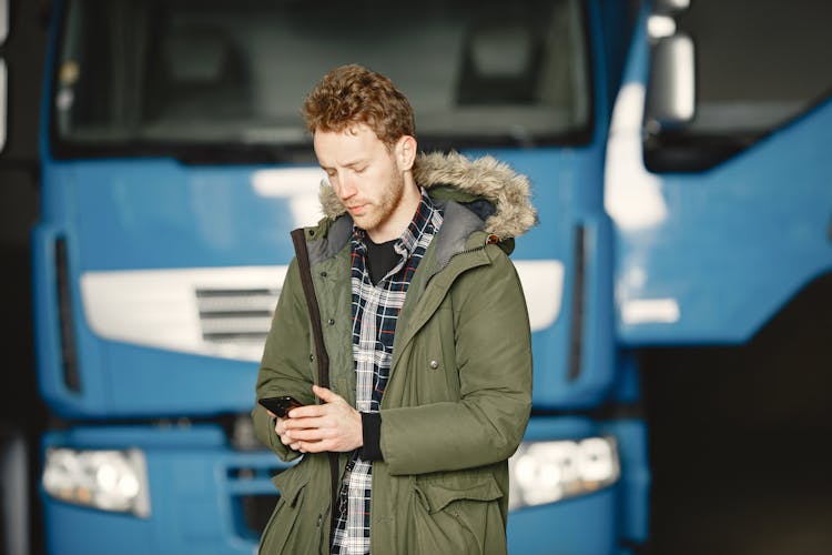 Man In Green Coat Holding Mobile Phone While Standing Beside Blue Truck