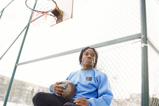 Young man sitting with a basketball on an outdoor court, wearing blue clothing.