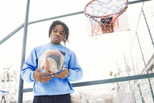 Young adult holding a basketball under an outdoor hoop in bright daylight.