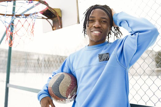 Smiling young man in blue sweatshirt holding a basketball outdoors next to a hoop.