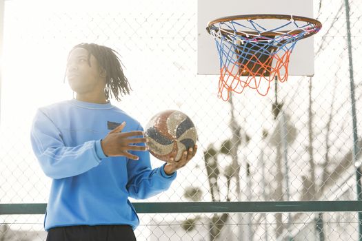 Young athlete casually holding basketball near hoop on outdoor court.