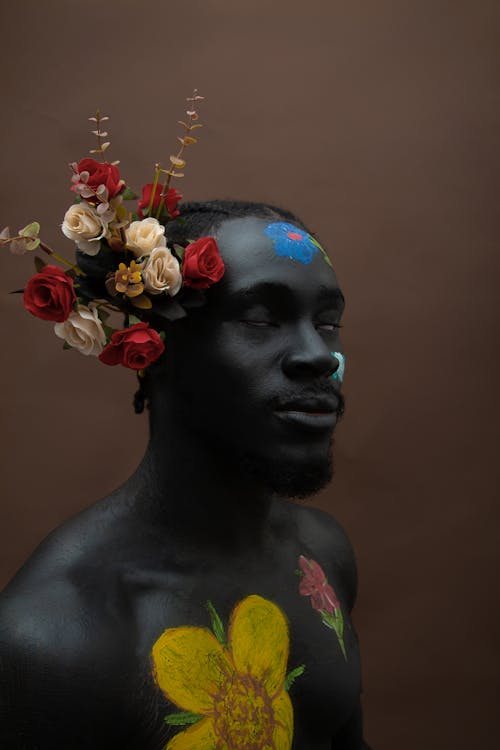 Free Emotionless young African American shirtless guy with bouquet with flowers behind ear with painted face and body covered in flowers on brown background Stock Photo