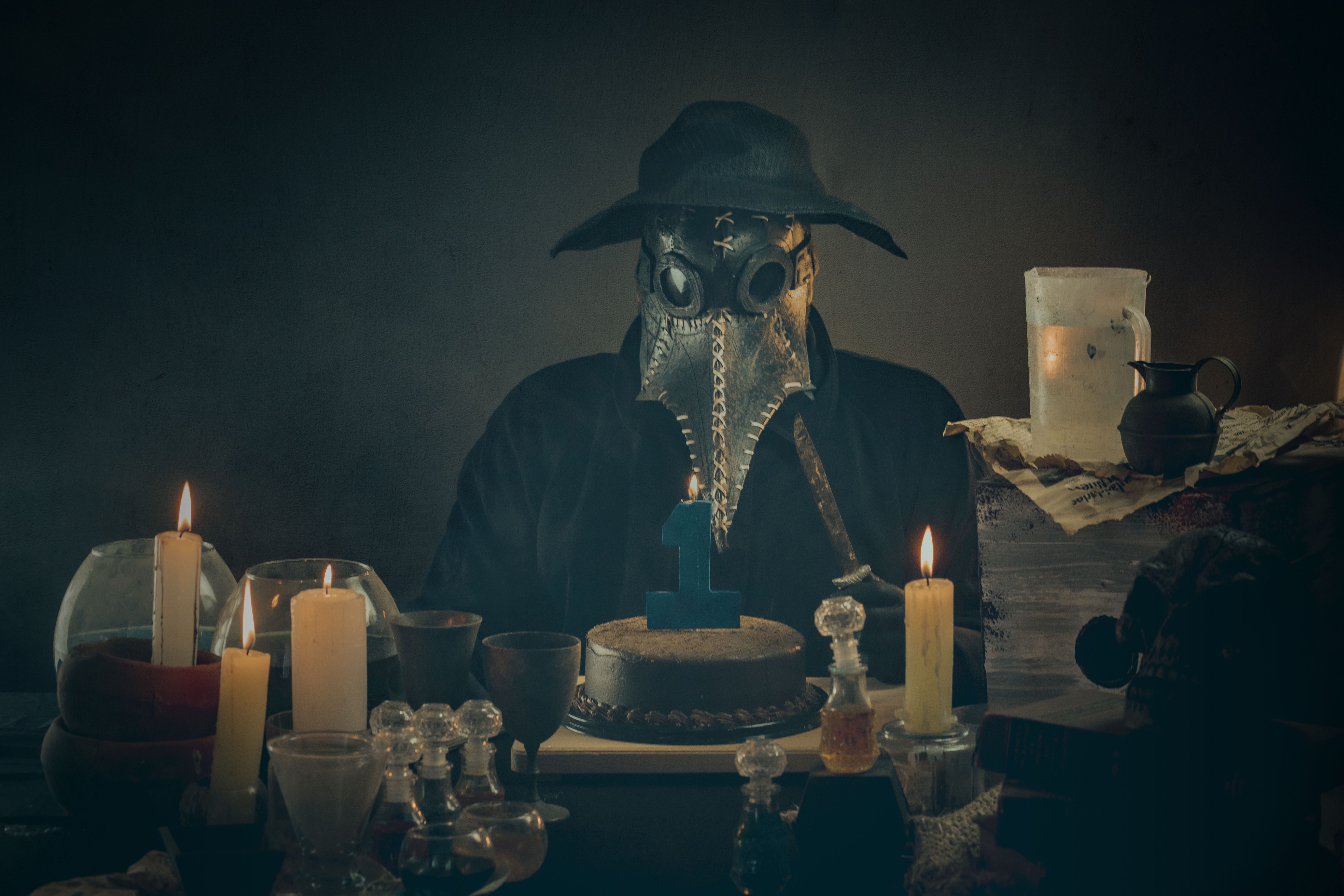 A masked figure resembling a plague doctor sits with candles and a cake in a dimly lit room.