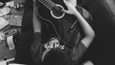 一个小男孩躺在室内的沙发上，弹着原声吉他，周围散落着书籍