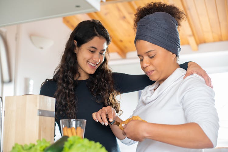 Woman Standing With Arm Around Girlfriend Making Breakfast