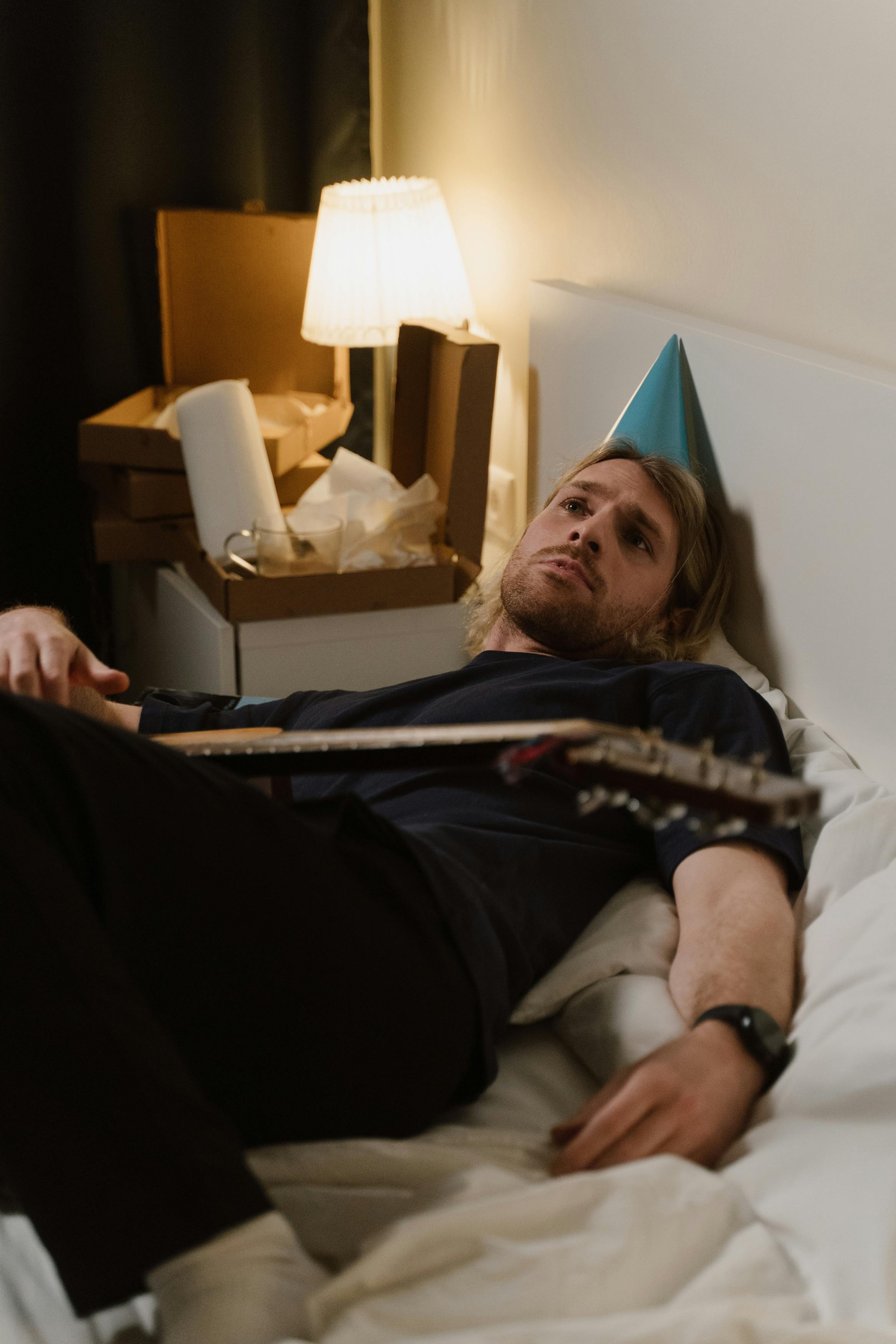 A musician with a guitar and party hat relaxing in a modern bedroom.