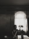 A Grayscale Photo of a Woman Sitting Near the Table