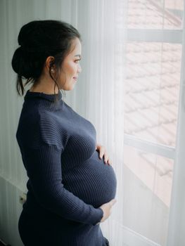 Pregnant woman in a blue sweater looking out a window, embracing motherhood. - Fotografer Sidoarjo