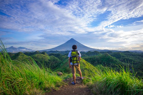 Free Man Wearing White Shirt, Brown Shorts, and Green Backpack Standing on Hill Stock Photo