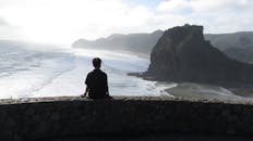 一个孤独的男孩坐在石墙上，凝视着奥克兰皮哈海滩的海洋与山脉