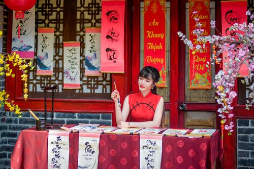 Free Asian woman in red cheongsam creating calligraphy art in traditional setting with festive decor. Stock Photo