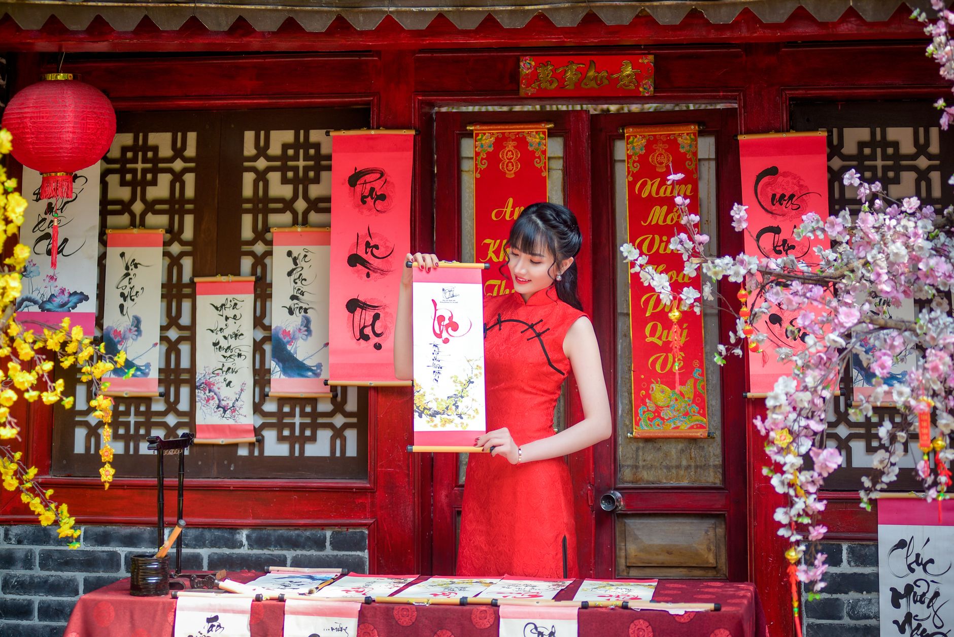 An adult woman in a red cheongsam holds traditional scrolls in a festive setting, symbolizing Asian culture.