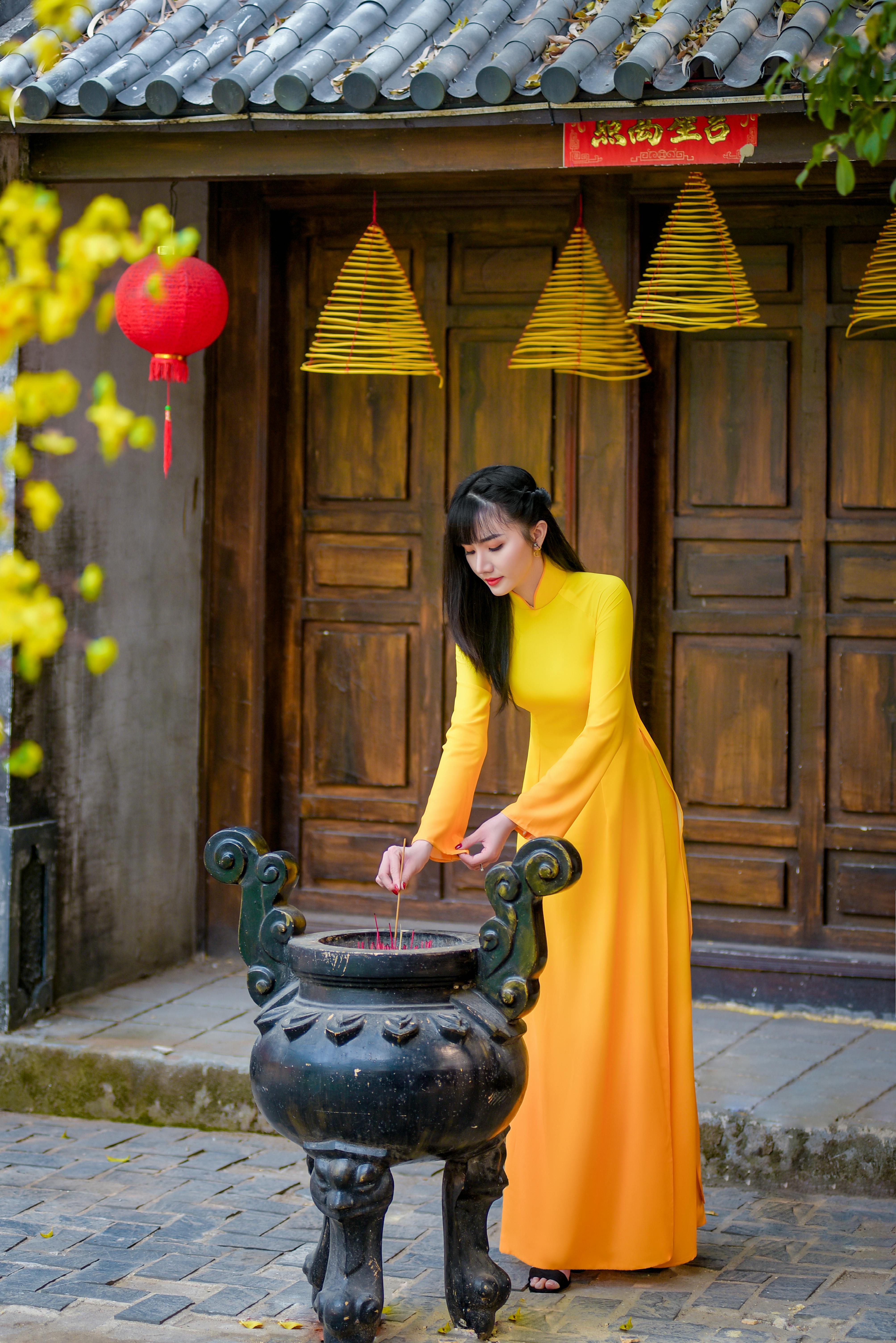 Asian woman in a vibrant yellow traditional dress lighting incense outdoors, focusing on cultural rituals.