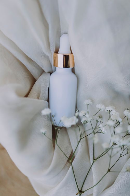 Free A white and gold dropper bottle elegantly placed with white flowers on soft fabric. Stock Photo