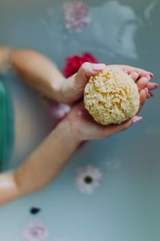 Hands holding a natural sea sponge over water surrounded by flowers for a relaxing spa experience.