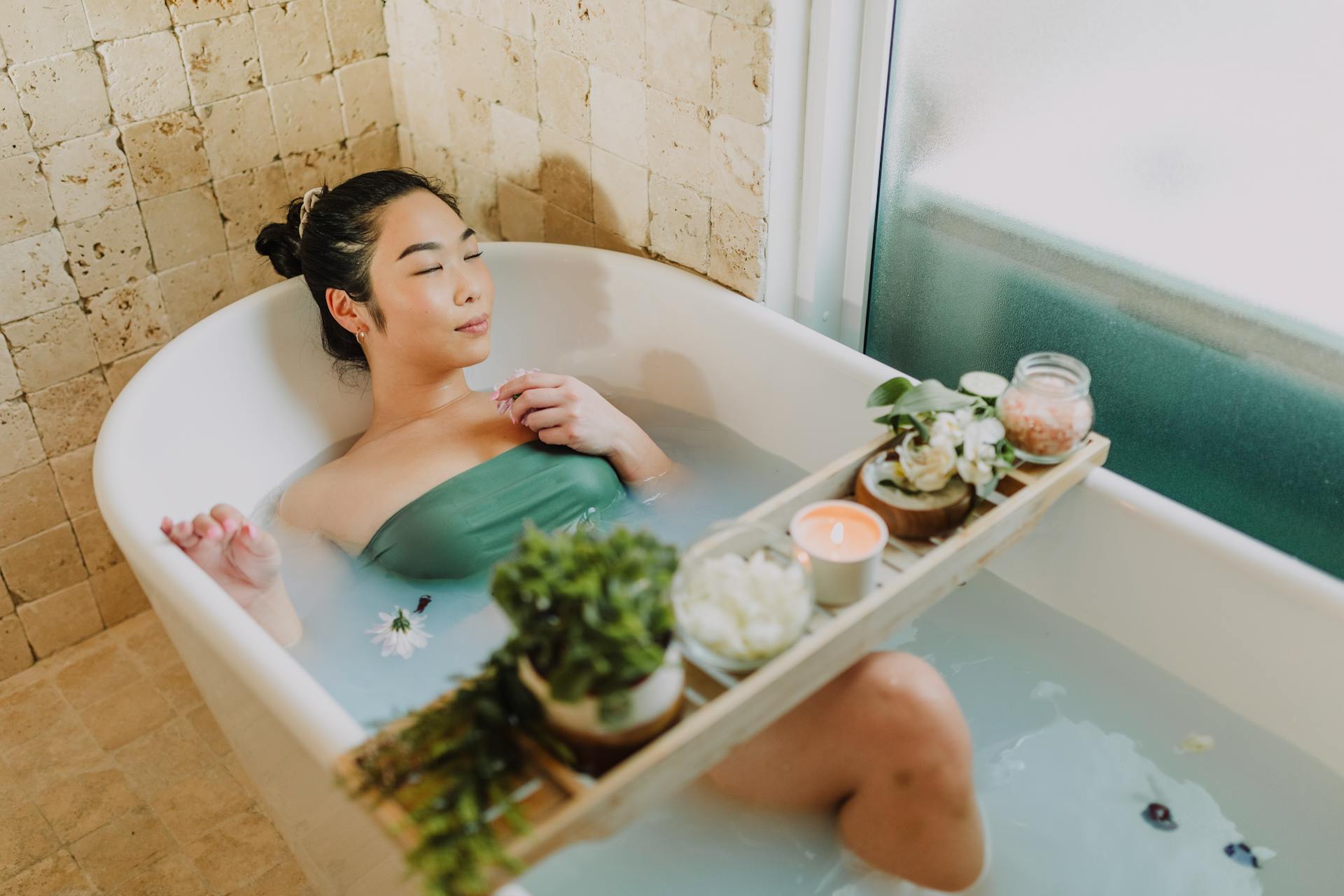 Asian woman relaxing in a luxurious bathtub with a bath board and candles, enjoying a serene spa moment.