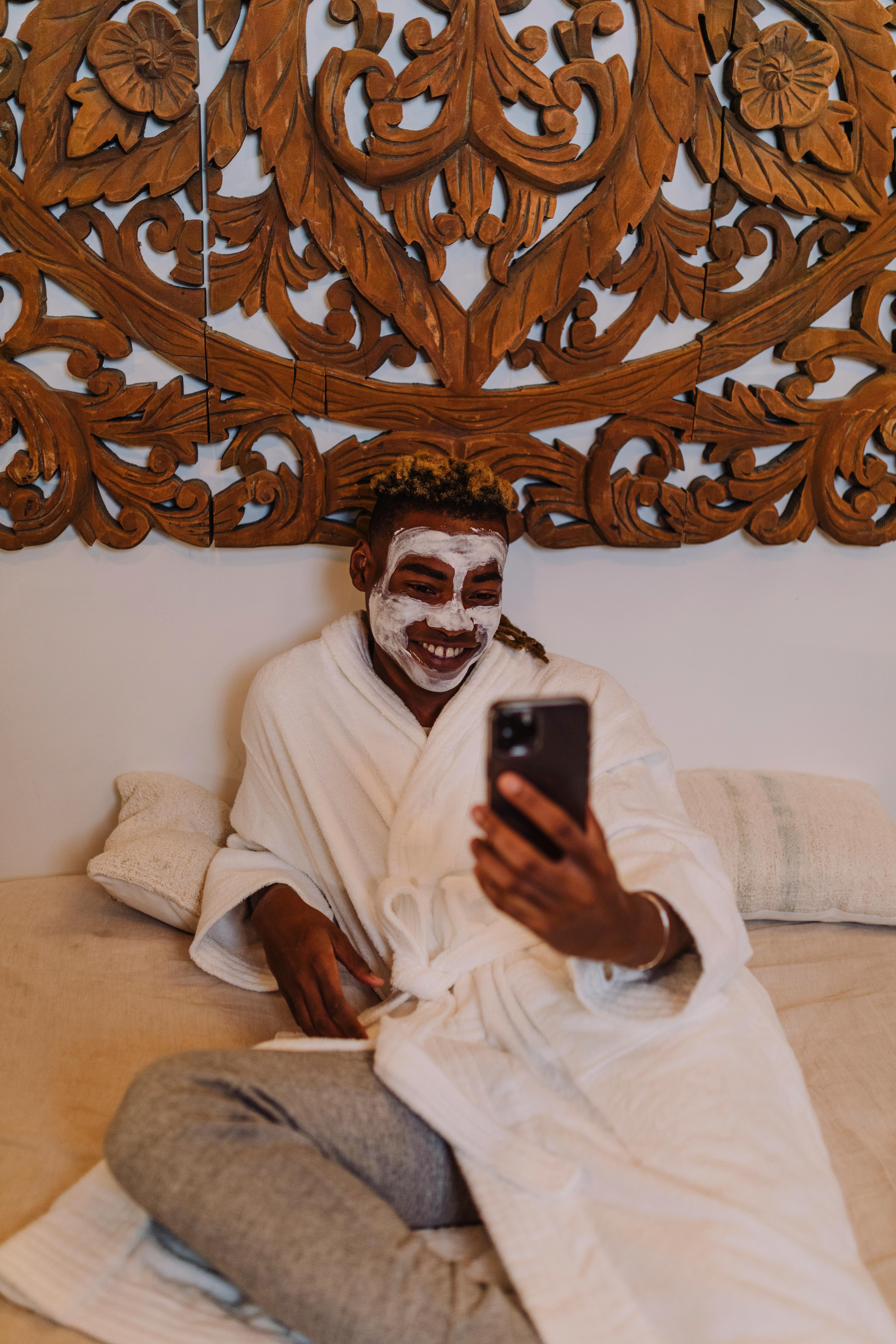 a man sitting on bed with cream on face holding a cellphone