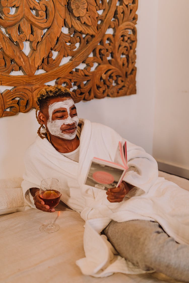 A Man Sitting On Bed With Cream On Face Holding A Book And A Glass Of Wine