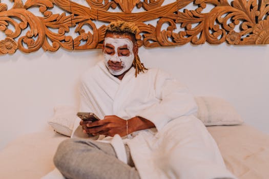 A man enjoys a relaxing spa day at home, applying a facemask while using his smartphone.