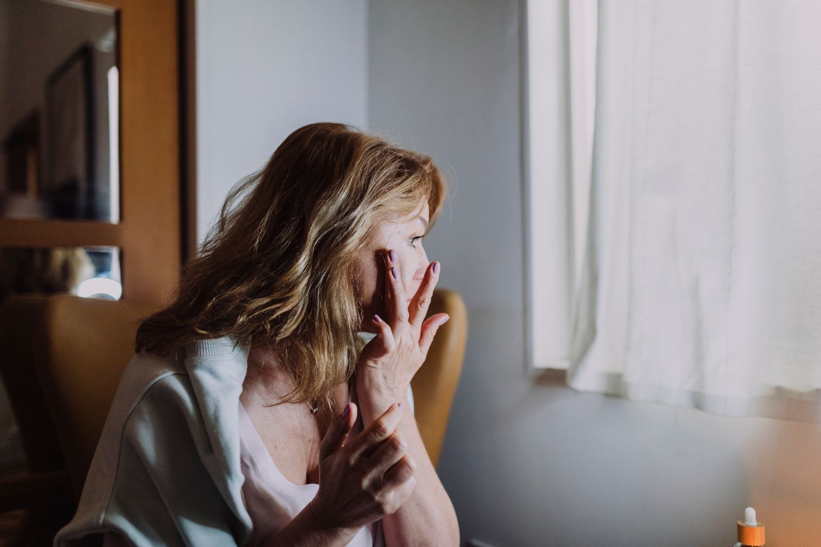 Woman sitting by a bright window gently touching her cheek while applying skincare serum