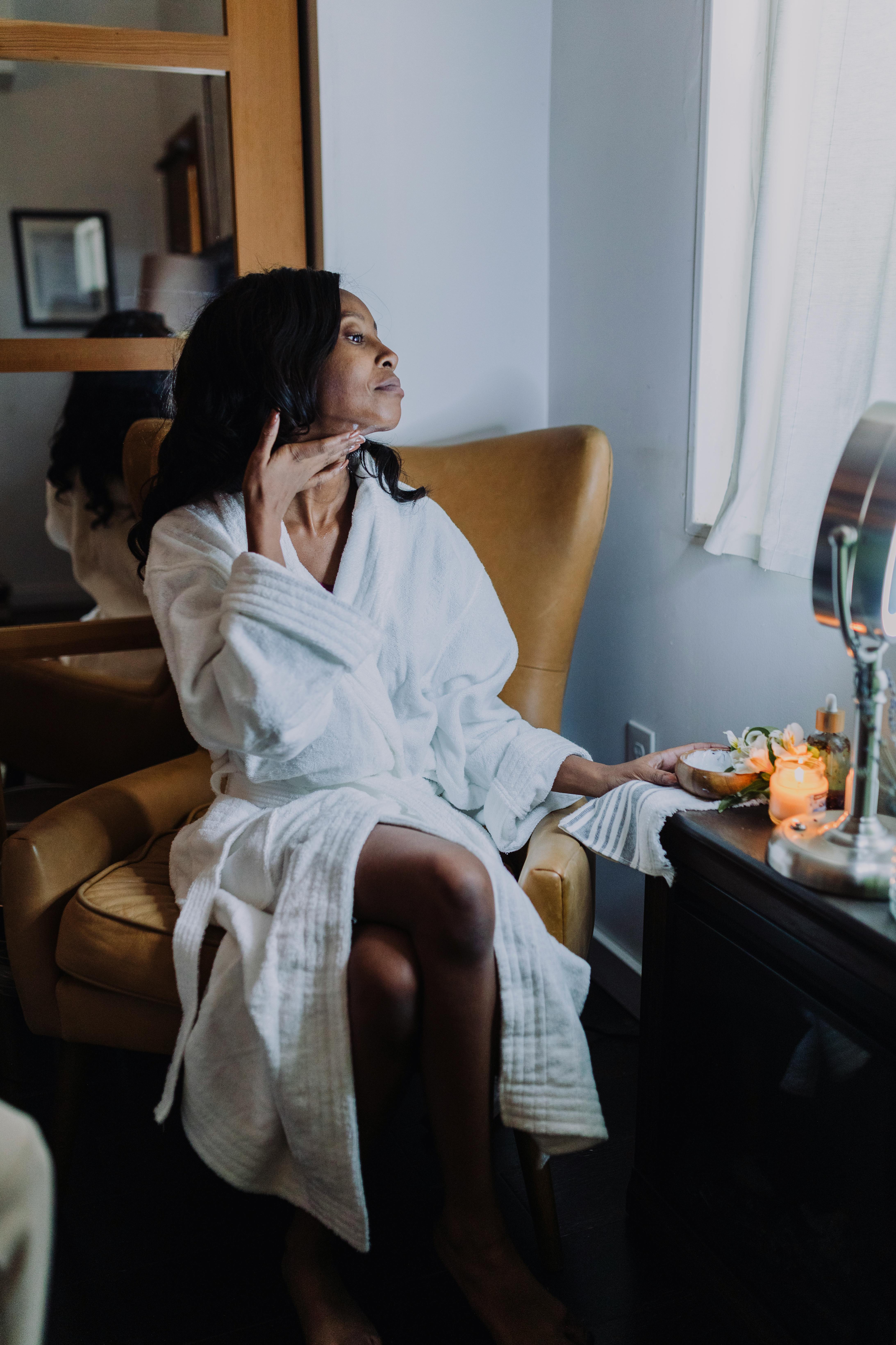 a woman in white robe sitting on chair applying cosmetic product under her chin