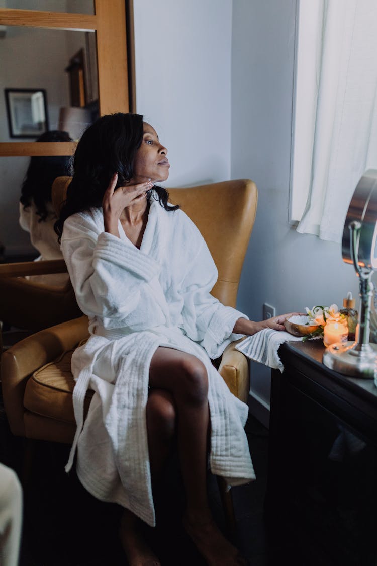 A Woman  In White Robe Sitting On Chair Applying Cosmetic Product Under Her Chin