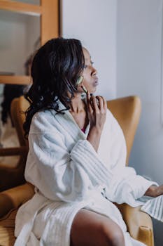 A woman in a robe using a jade roller for skincare, sitting indoors in a cozy setting.