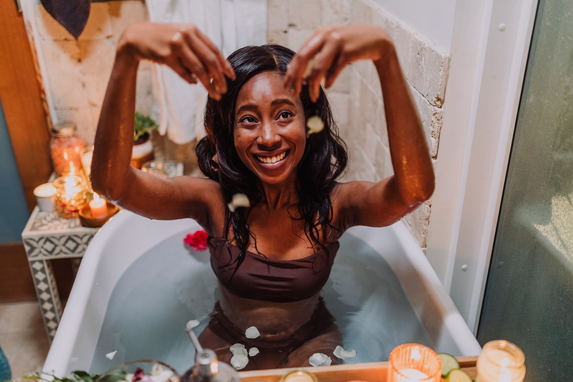 A smiling woman enjoys a relaxing bath surrounded by candles and flower petals, embodying self-care and relaxation.